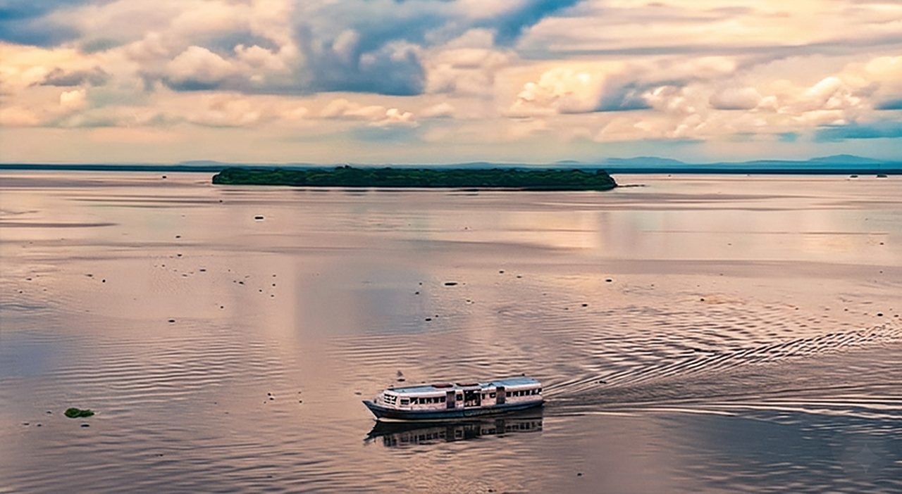 A passenger service boat leaving a wake on a wide, calm lake under a golden cloudy sky, with a small wooded island on the horizon