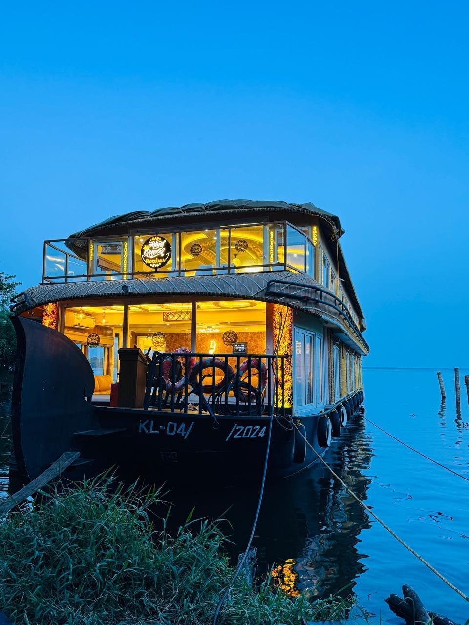 Royal Queen Roxxlana, a two-deck houseboat, lit up in gold at dusk beside tall grass on the shore