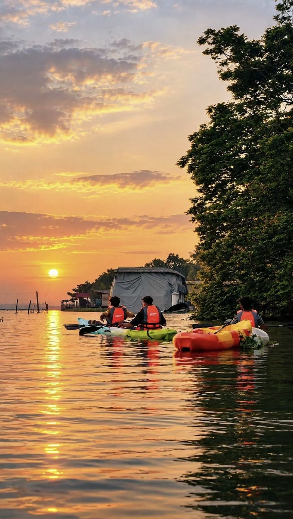 Three kayakers in orange life jackets paddling toward the setting sun on calm, golden water